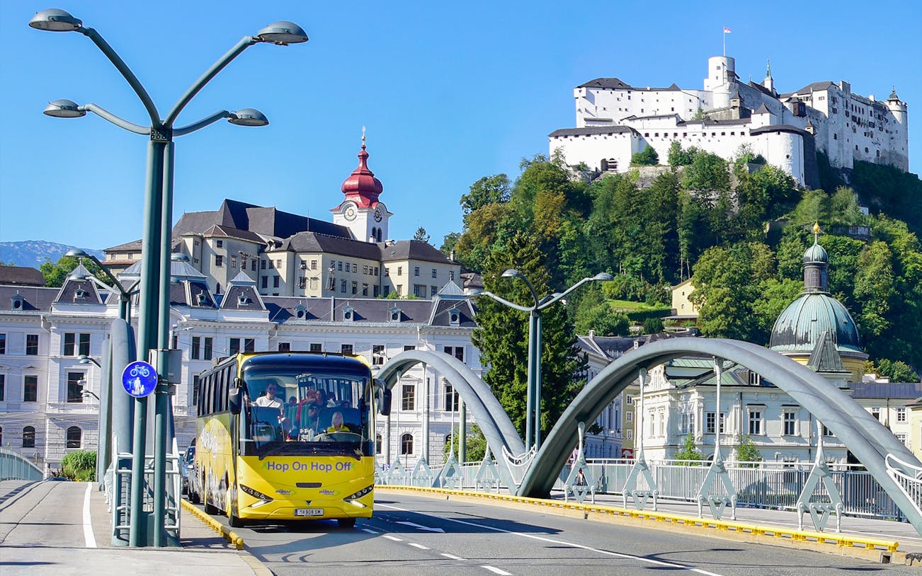 Hop On Hop Off bus crossing a bridge in Salzburg with Hohensalzburg Fortress in the background.