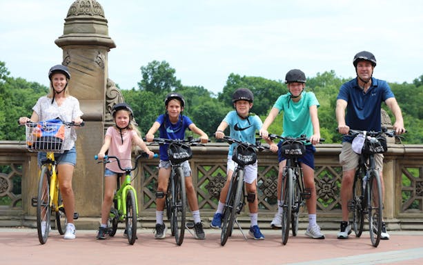Family biking in Central Park, New York City, with rental bikes on a scenic bridge.