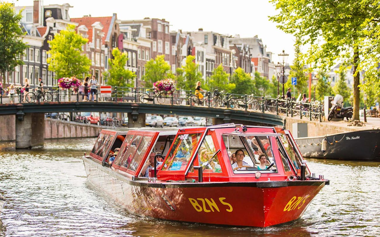 Red canal boat on Amsterdam waterway with tourists, bridge, and historic buildings in view.