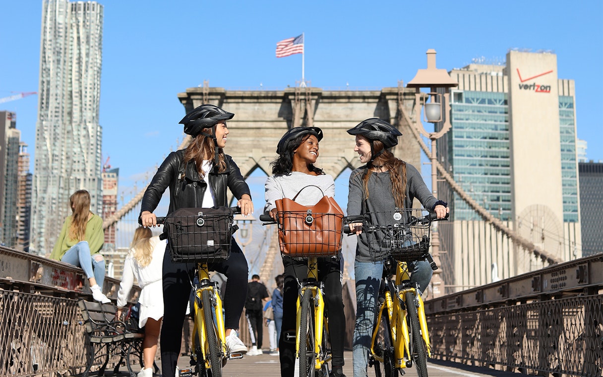 Cyclists on Brooklyn Bridge with city skyline in background.