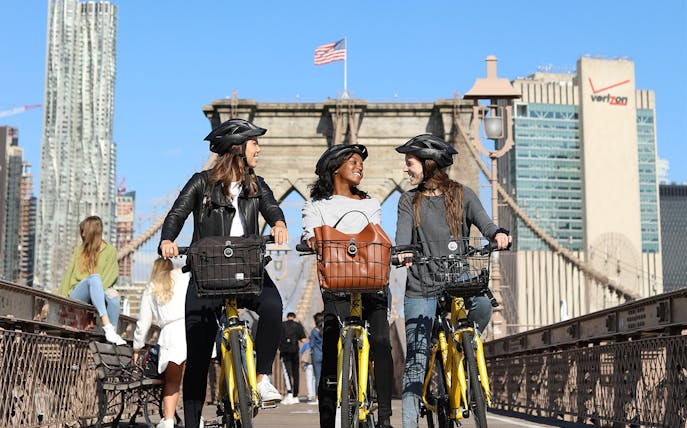 Cyclists on Brooklyn Bridge with city skyline in background.