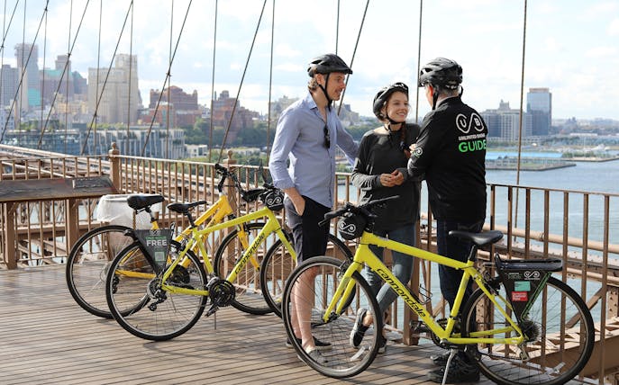 Cyclists with a guide on Brooklyn Bridge during a bike tour, New York City skyline in background.
