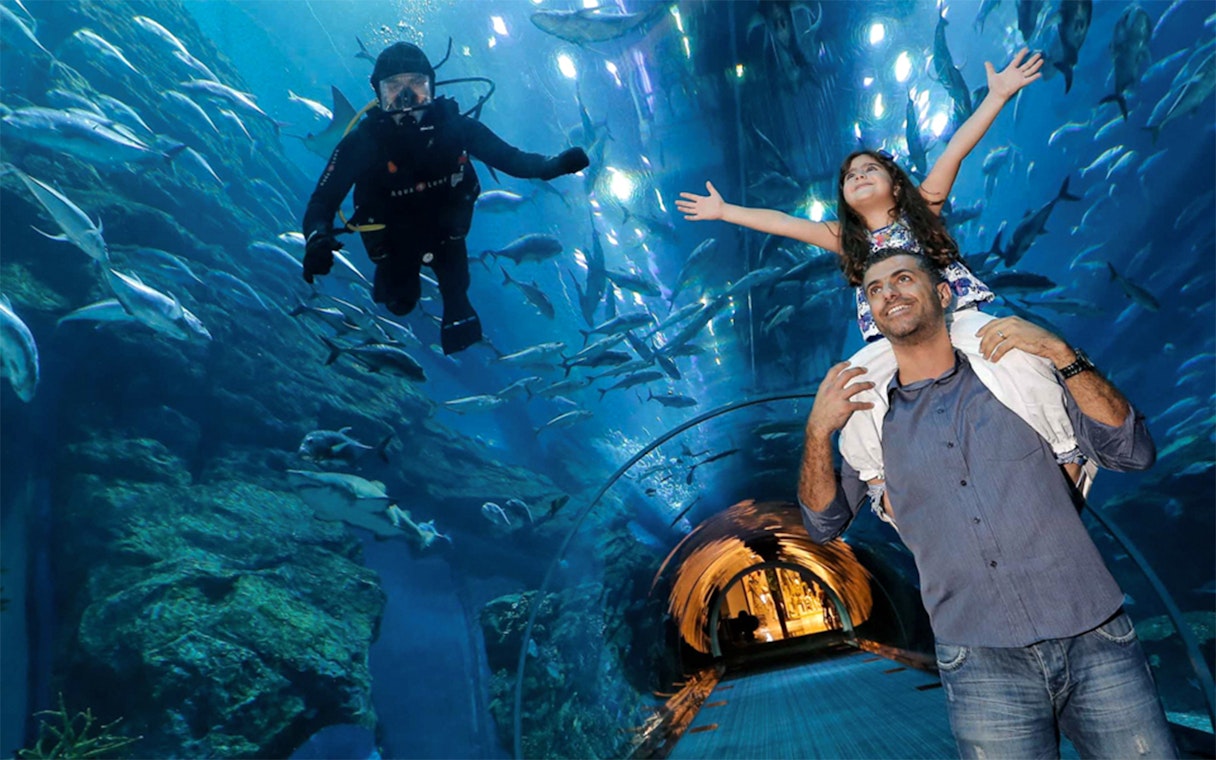 Father and daughter in Dubai Aquarium tunnel with diver and fish overhead.
