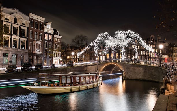 Canal cruise boat under illuminated bridge during Amsterdam Light Festival.