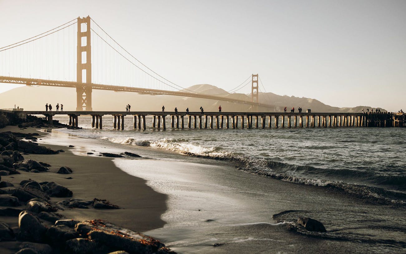 Golden Gate Bridge view from a beach in San Francisco, with people walking on a pier.