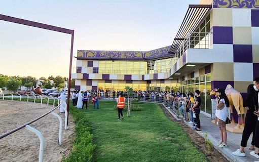 Spectators gather at Dubai Royal Camel Racing venue, with track and modern building in view.