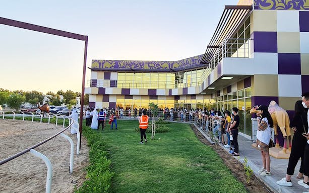 Spectators gather at Dubai Royal Camel Racing venue, with track and modern building in view.