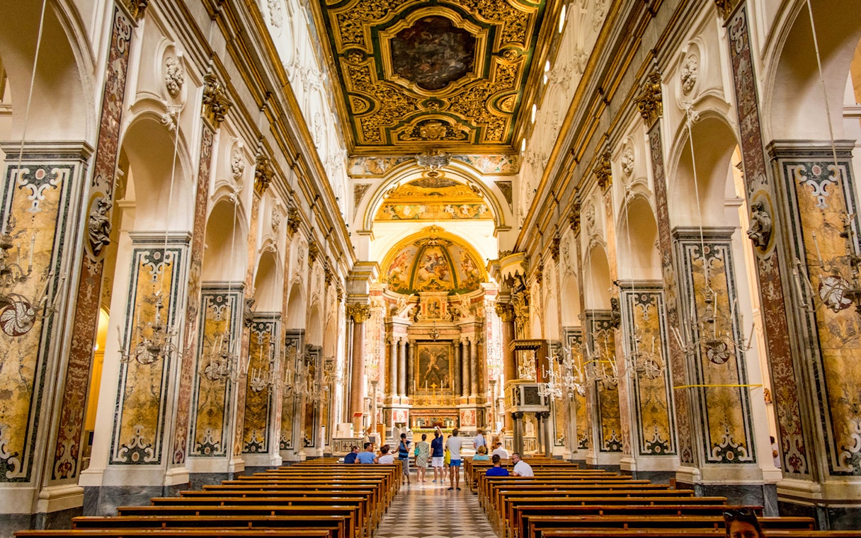 Interior of a historic church in Amalfi with ornate ceiling and visitors exploring.