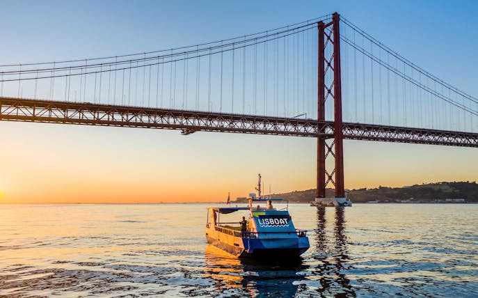 Lisbon tour boat under 25 de Abril Bridge at sunset.