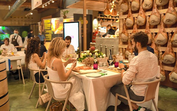 Guests dining at FICO Eataly World, Bologna, surrounded by hanging cured meats.