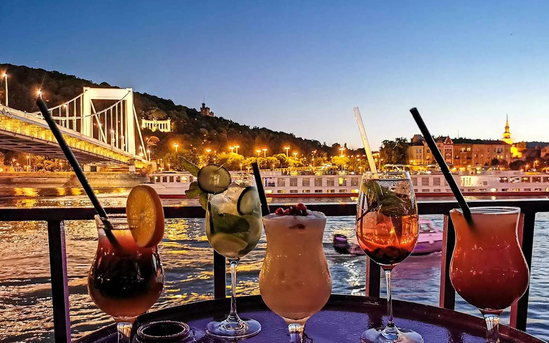 Cocktails on a table with Budapest's illuminated skyline and Danube River at sunset.