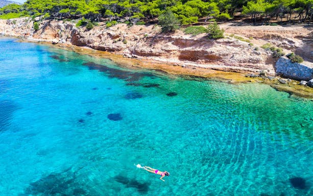 Person swimming in clear blue waters near a rocky coastline in Athens.