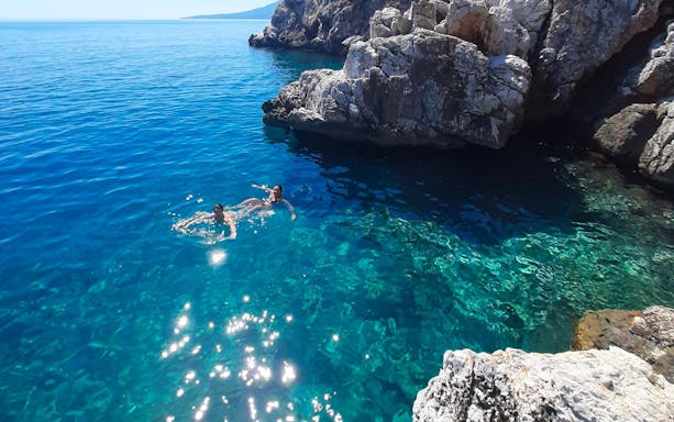 Swimmers in clear blue water near rocky coast, Athens cruise tour.