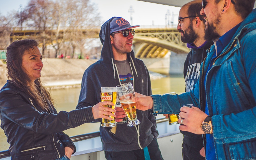 Group enjoying drinks on a Budapest party boat cruise near a bridge.
