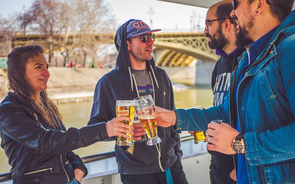 Group enjoying drinks on a Budapest party boat cruise near a bridge.