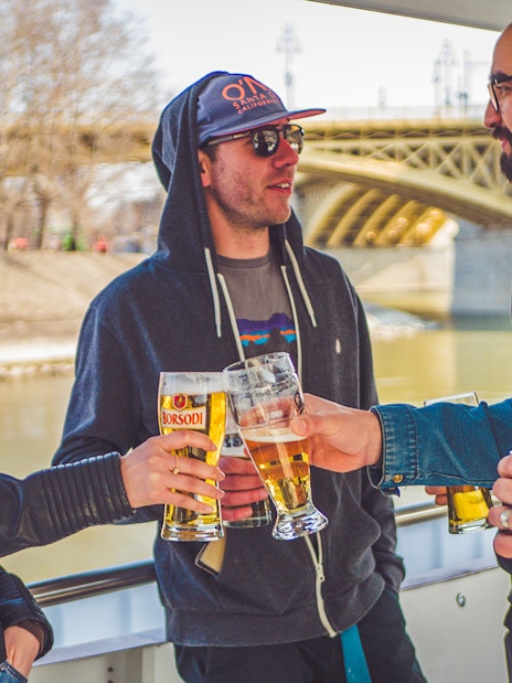 Group enjoying drinks on a Budapest party boat cruise near a bridge.