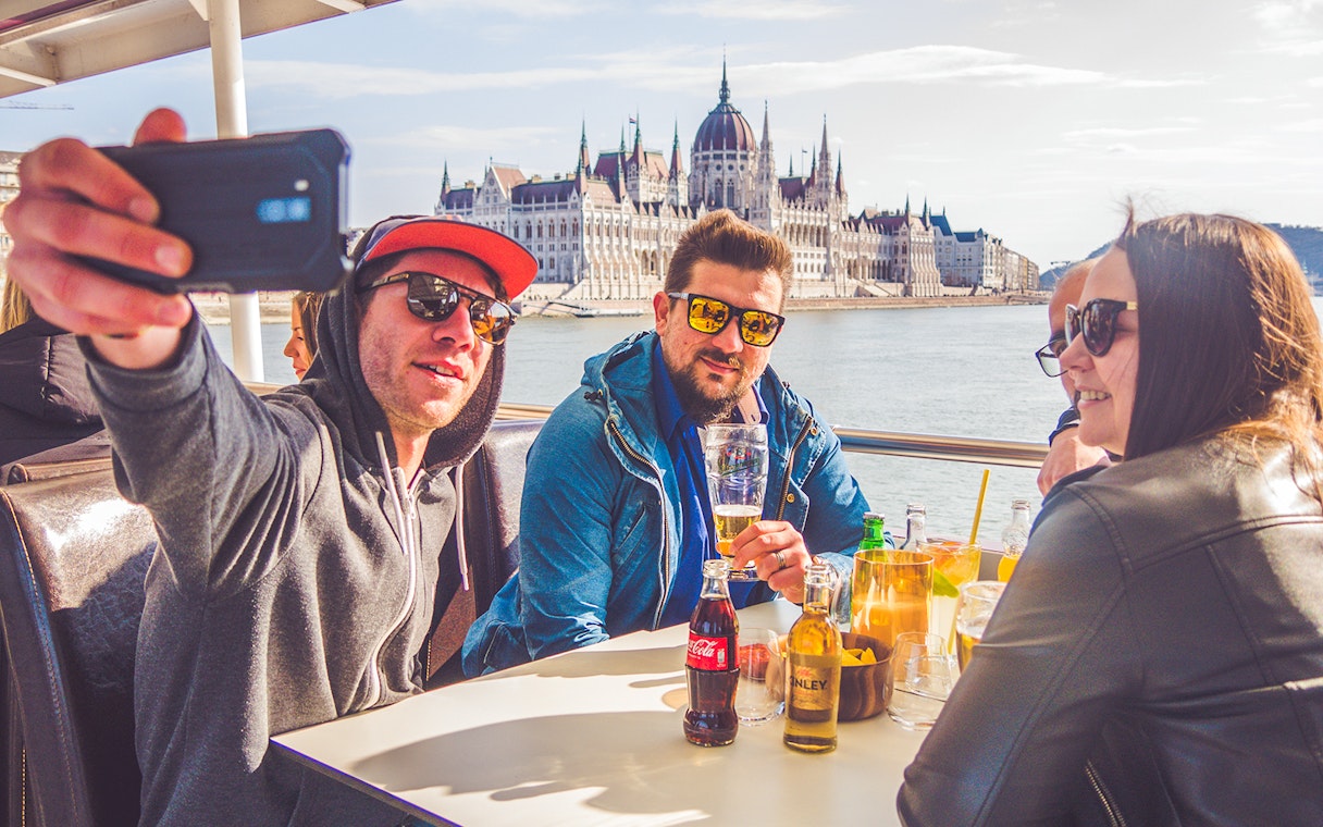 Group enjoying drinks on Budapest party boat cruise with Parliament building in background.