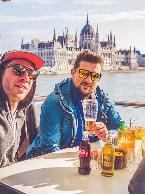 Group enjoying drinks on Budapest party boat cruise with Parliament building in background.