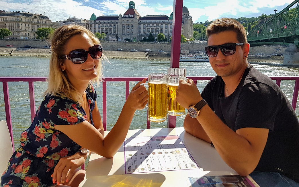 Couple enjoying beers on a Budapest party boat cruise with cityscape view.