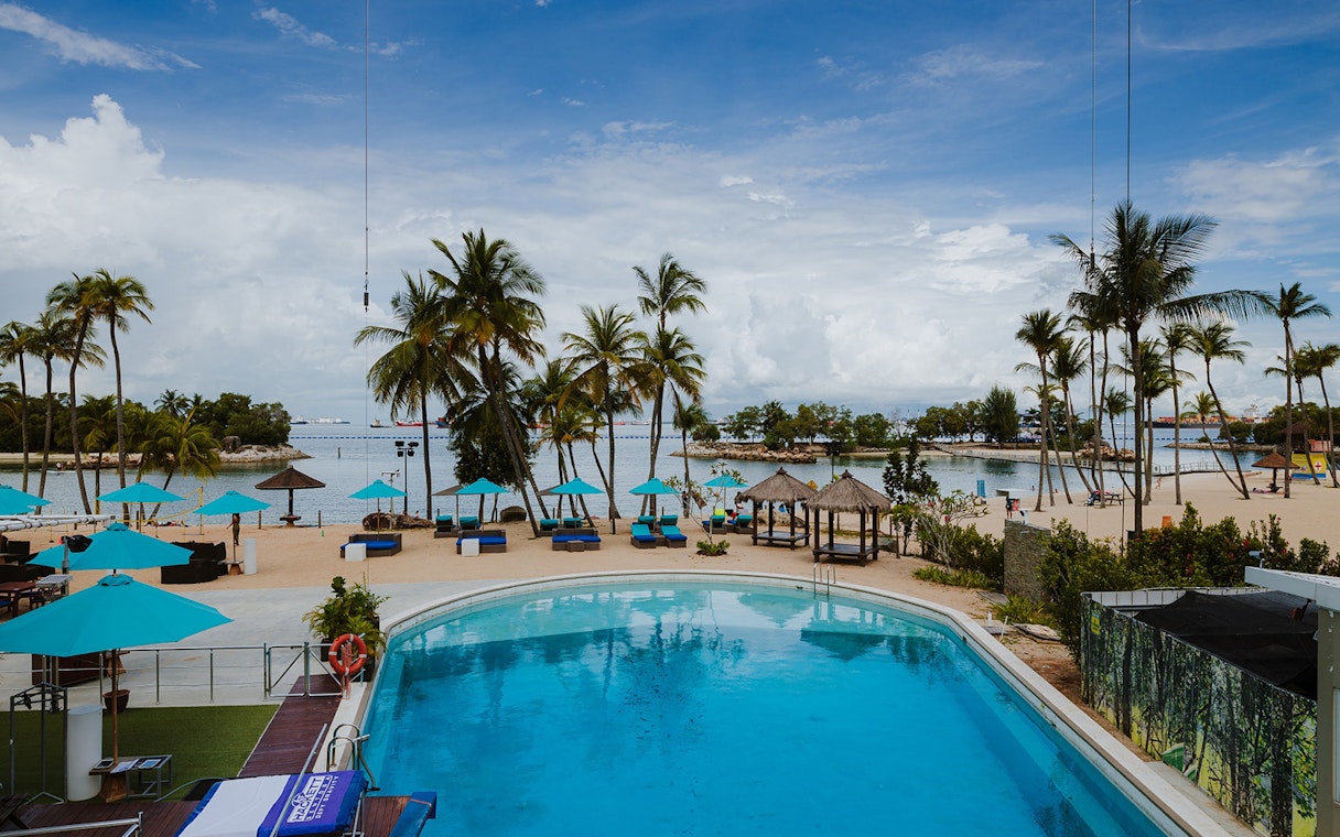 Sentosa beach view with poolside dining area and palm trees.