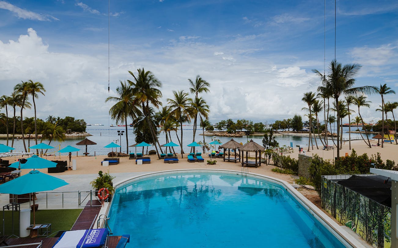 Sentosa beach view with poolside dining area and palm trees.