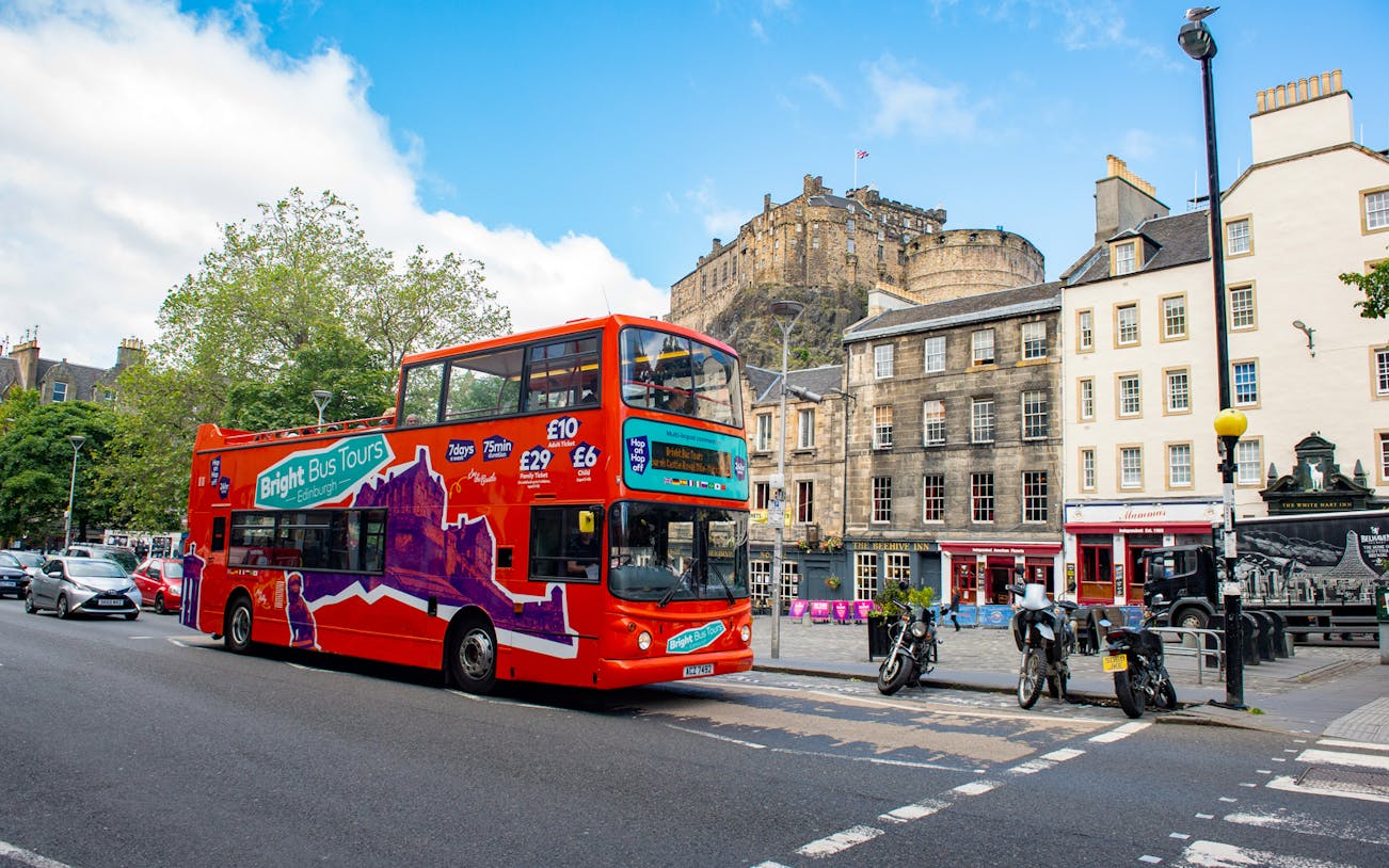 Red double-decker bus on Edinburgh street with castle in background, part of Bright Bus City Tours.