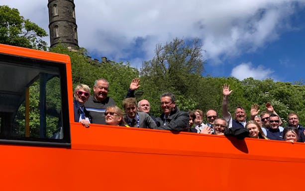 Open-top orange bus with tourists waving, passing a historic tower on a Bright Bus City Tour.