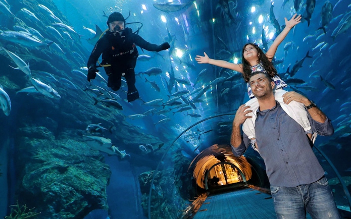 Father and daughter in Dubai Aquarium tunnel with diver and fish overhead.