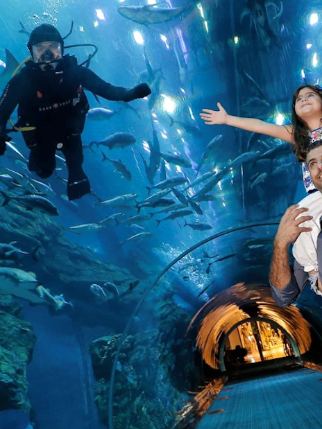 Father and daughter in Dubai Aquarium tunnel with diver and fish overhead.