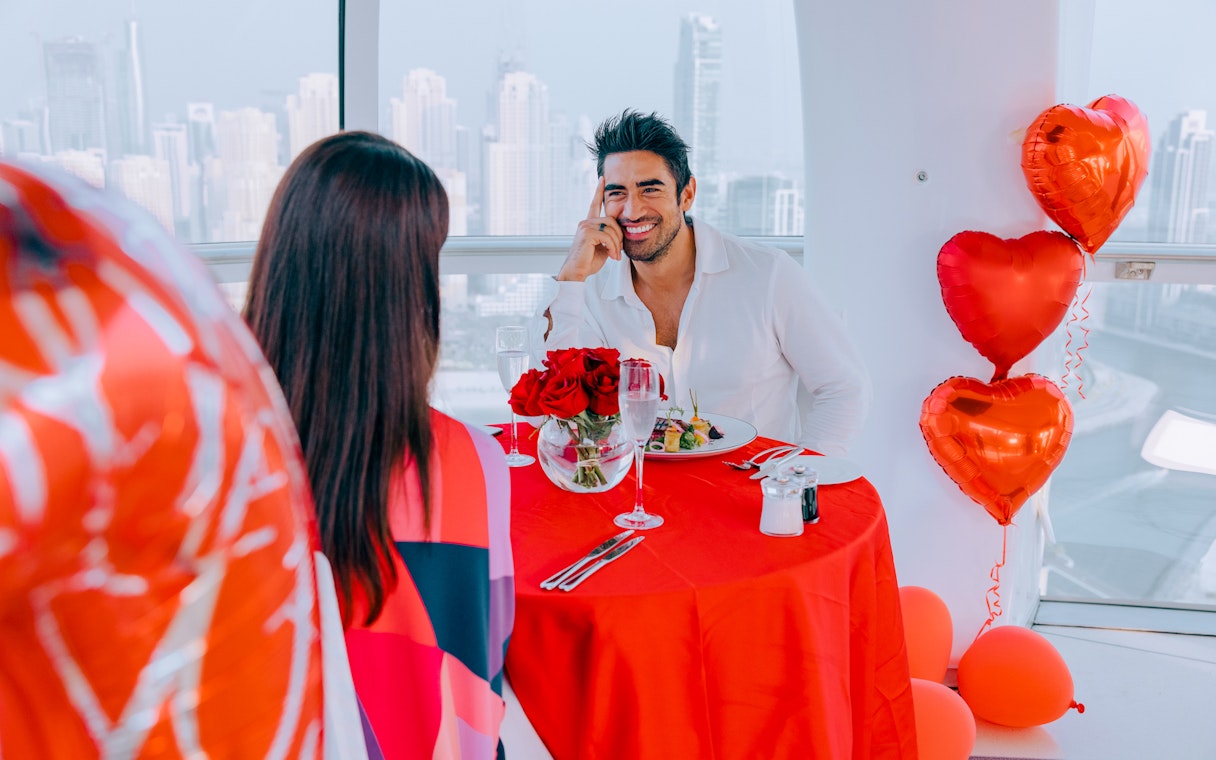 Couple dining with city view at Ain Dubai, red balloons and roses on table.