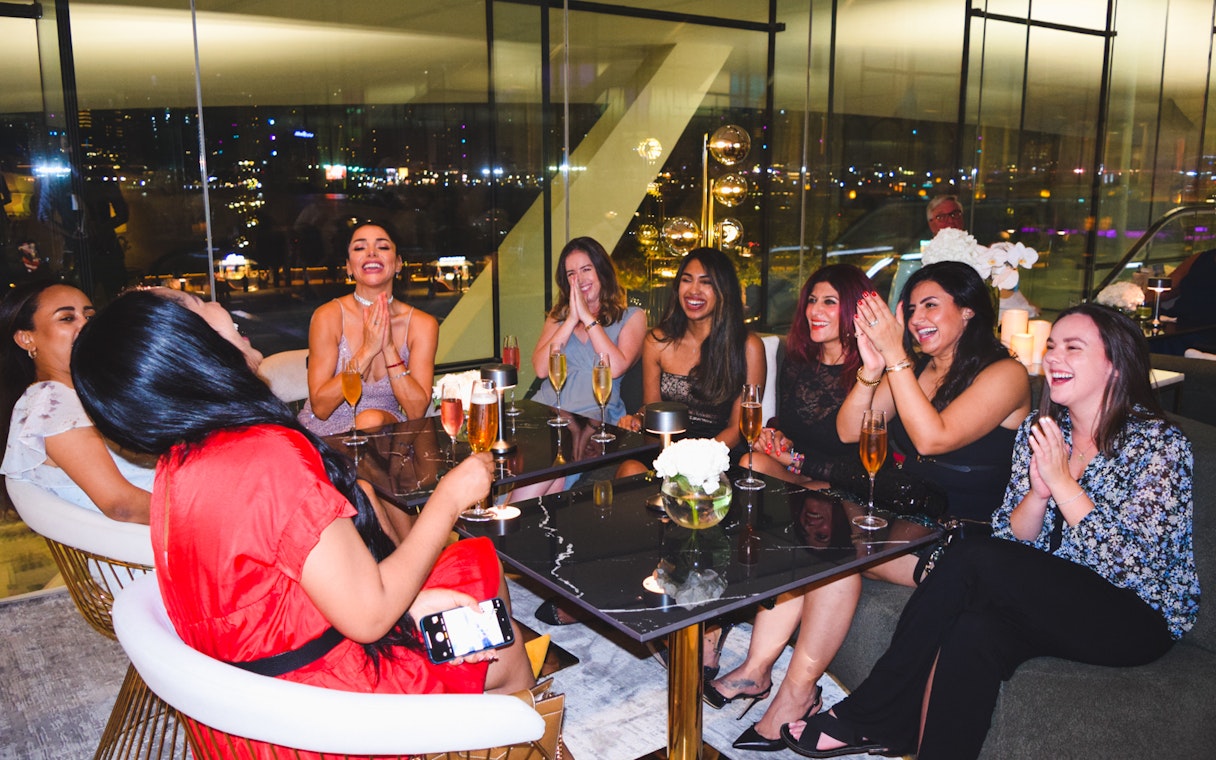 Group of friends enjoying drinks at a Dubai lounge with city lights in the background.