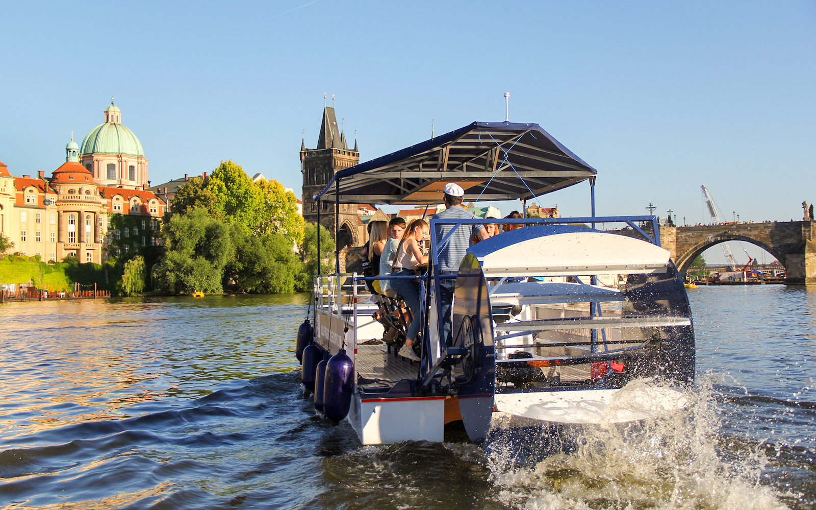 Cycle boat on Vltava River near Charles Bridge, Prague, with passengers enjoying the view.