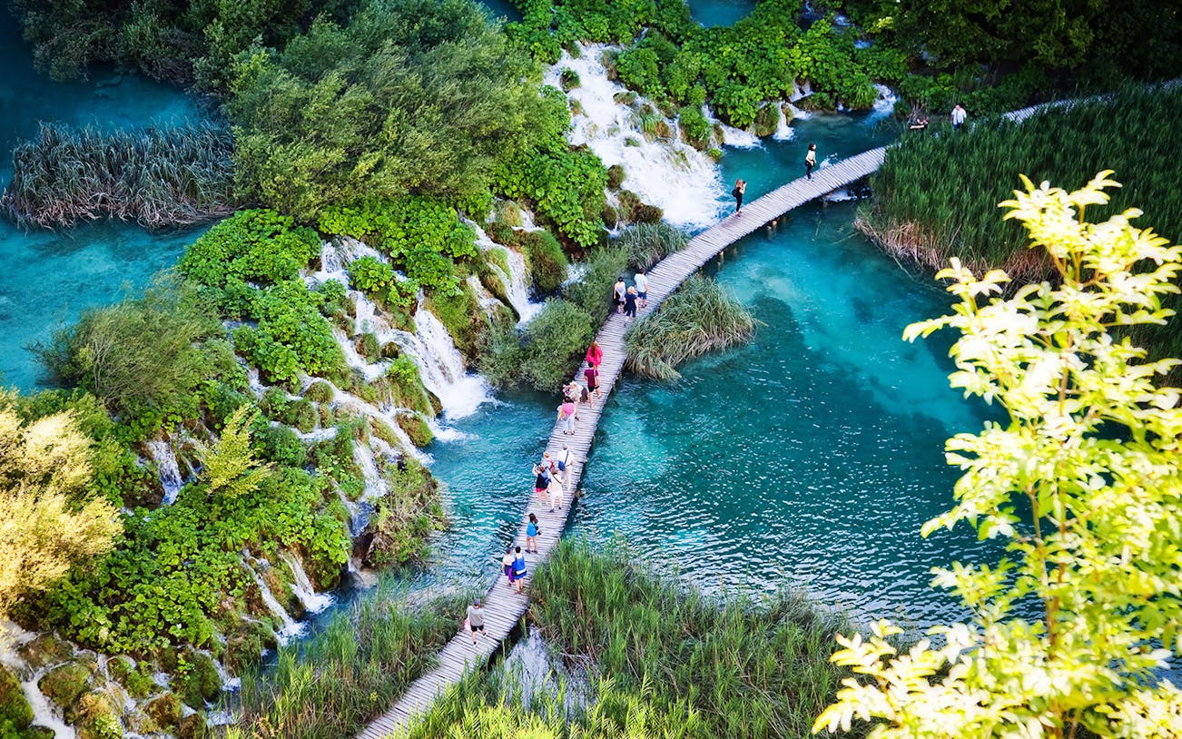 Visitors walking on a wooden path over turquoise lakes and waterfalls in Plitvice Lakes National Park, Croatia.