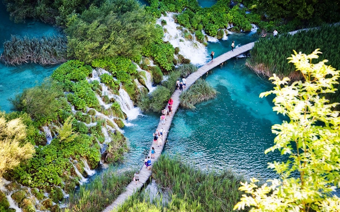 Visitors walking on a wooden path over turquoise lakes and waterfalls at Plitvice Lakes National Park.