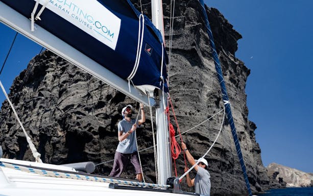 Sailors adjusting sails on a yacht near rocky cliffs during Caldera Day Cruise.