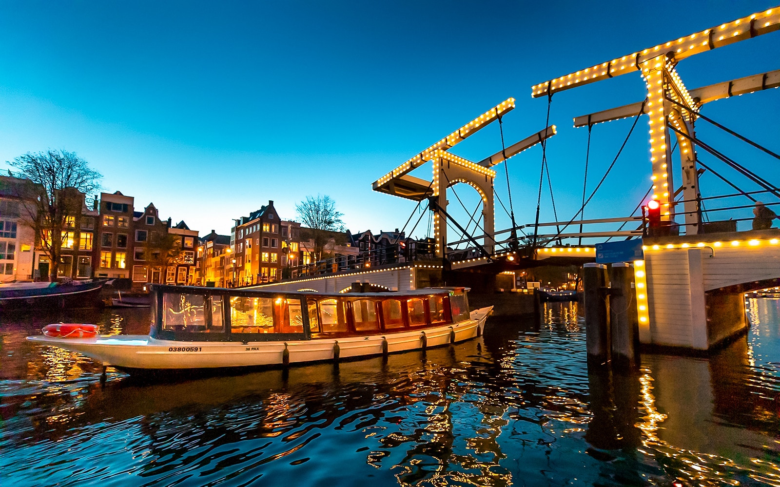 Boat passing under illuminated Skinny Bridge in Amsterdam at dusk.