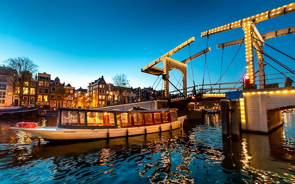 Boat passing under illuminated Skinny Bridge in Amsterdam at dusk.