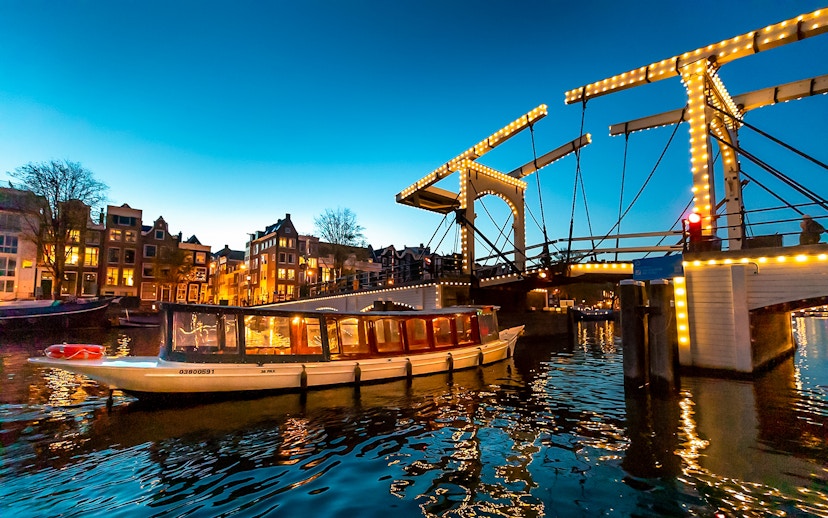 Boat passing under illuminated Skinny Bridge in Amsterdam at dusk.
