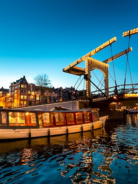 Boat passing under illuminated Skinny Bridge in Amsterdam at dusk.