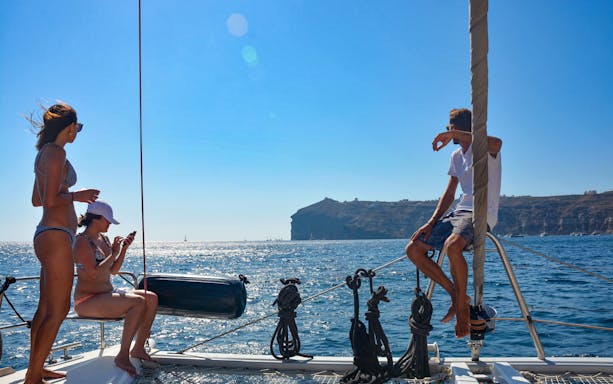 Sailing catamaran with people enjoying views of Santorini cliffs and sea.
