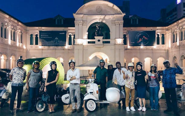 Group of people with vintage Vespas in front of Singapore Art Museum at night.