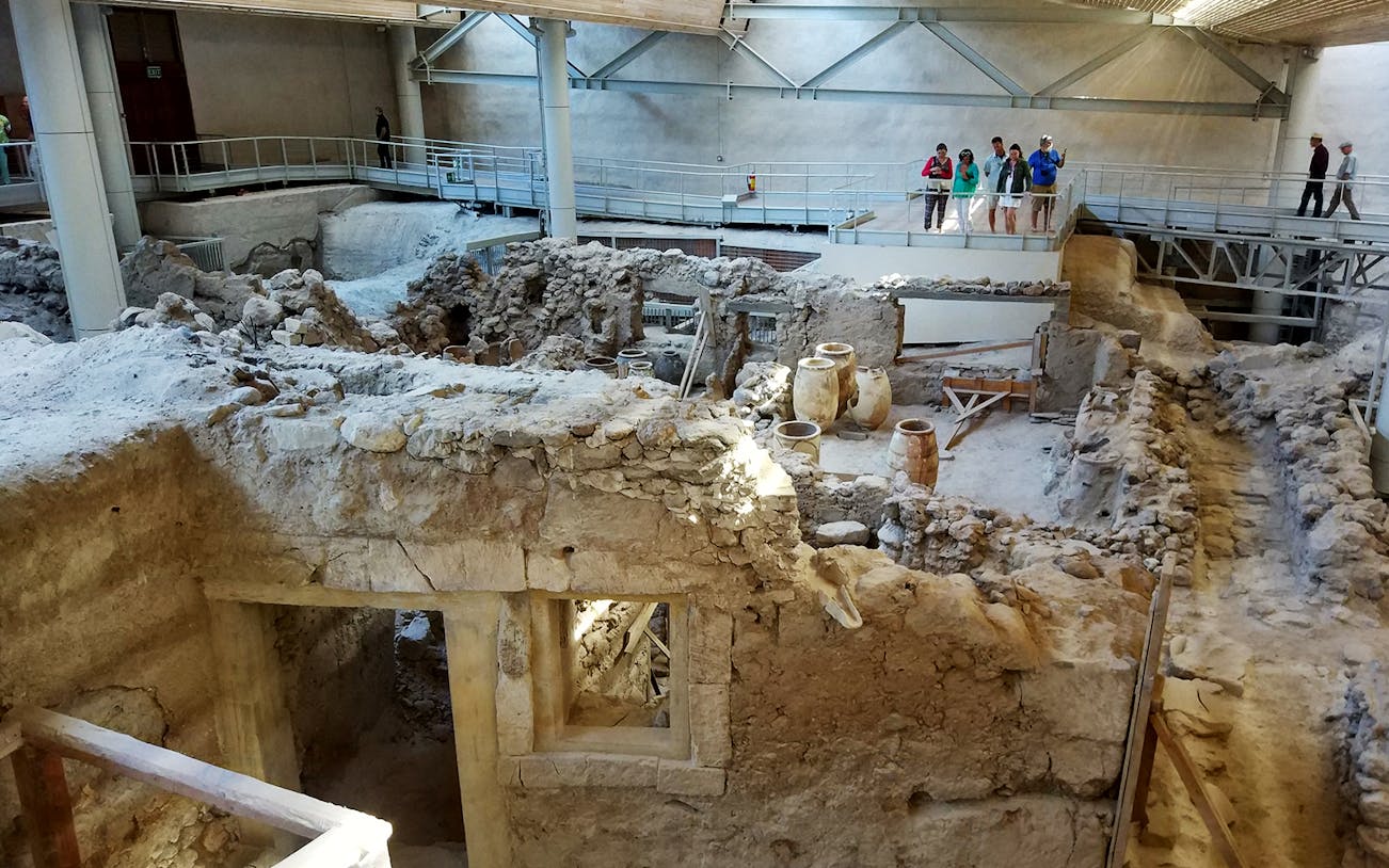 Akrotiri ruins with ancient pottery and tourists on a walkway during a guided tour.