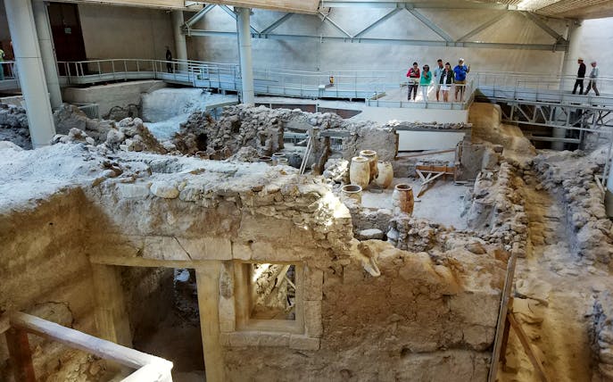 Akrotiri ruins with ancient pottery and tourists on a walkway during a guided tour.