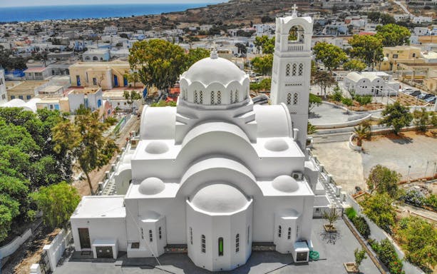 White church with domed roof and bell tower in Santorini, Greece, surrounded by trees and buildings.