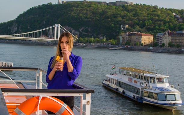 Danube cruise with passenger enjoying drink, Budapest in background.