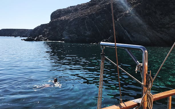 Sailboat near rocky coast with swimmer, Delos and Rhenia islands cruise.