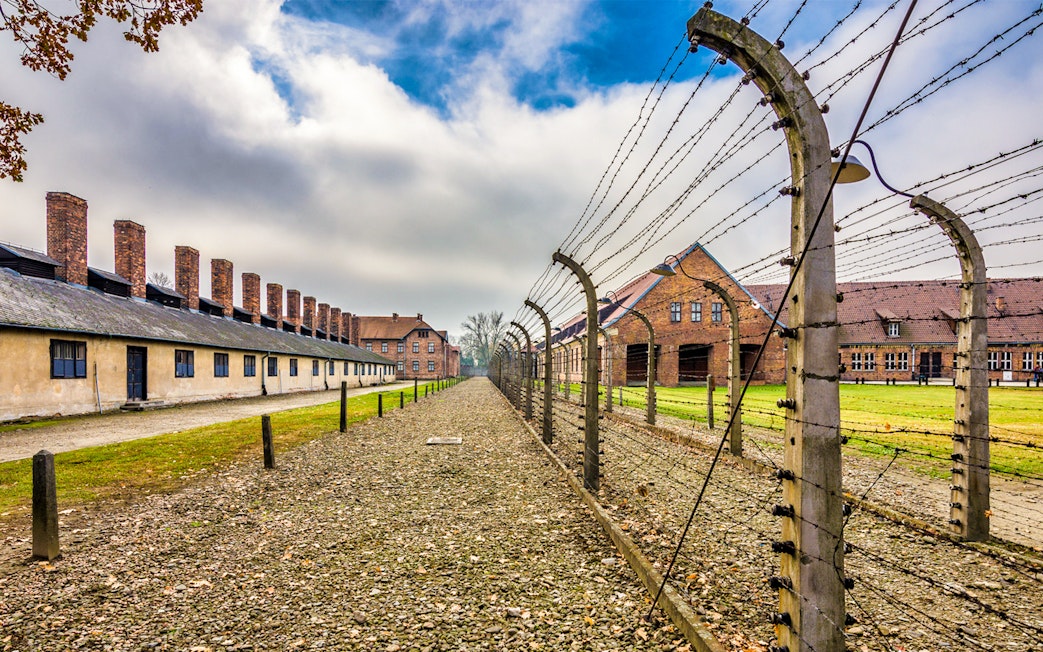 Auschwitz-Birkenau concentration camp buildings and barbed wire fence under cloudy sky.