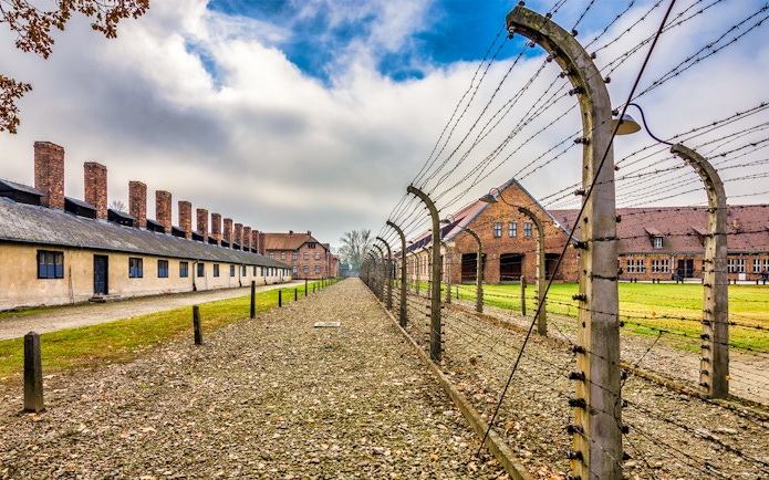 Auschwitz-Birkenau concentration camp buildings and barbed wire fence under cloudy sky.