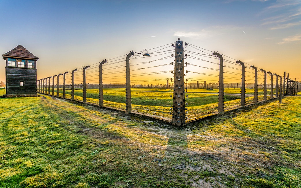 Barbed wire fence and guard tower at Auschwitz-Birkenau during sunset.