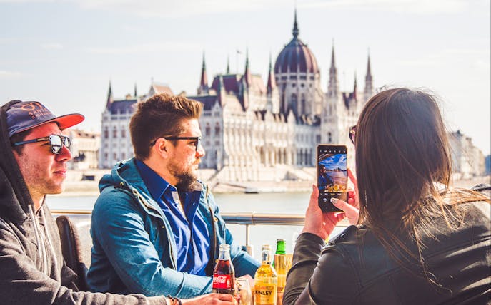 Tourists enjoying drinks on a Budapest sightseeing cruise with the Hungarian Parliament in view.